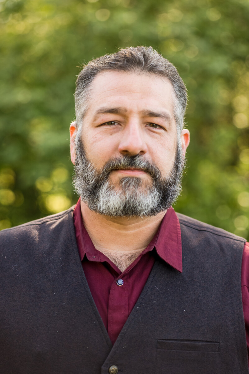 Matt with grey and brown beard and mustache and short brown hair with maroon colored collard button up shirt with a brown sweater over the top of the shirt.